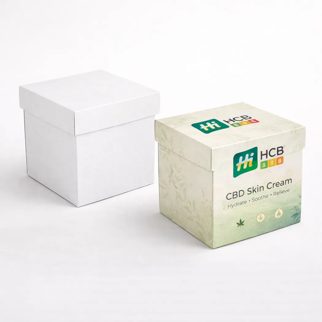 Two square cardboard boxes- one plain white and one featuring green botanical branding for CBD cream- class=